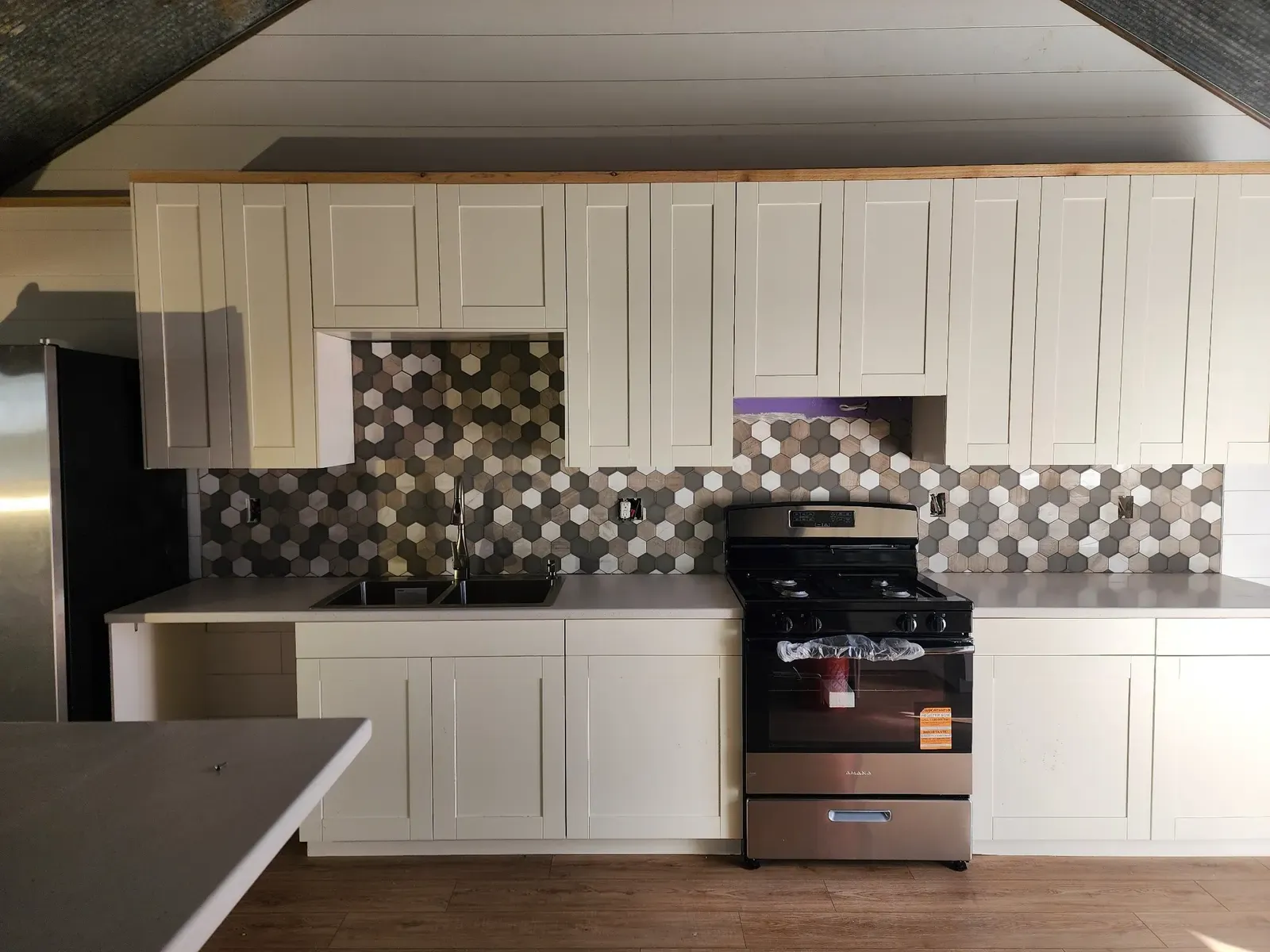 Wide view of the finished cabin kitchen with white cabinetry, hexagon tile backsplash, sink, black stove, and stainless steel refrigerator