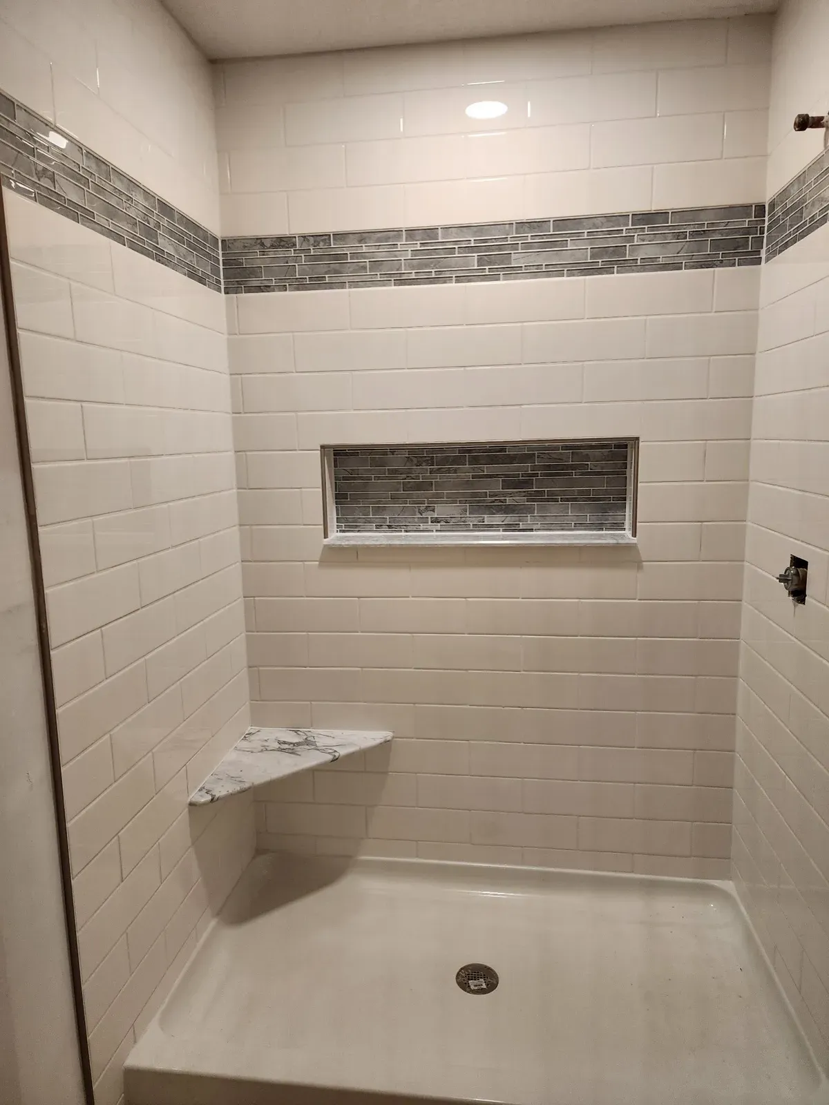Completed walk-in shower with white subway tile, mosaic accent band, recessed niche, and marble corner bench